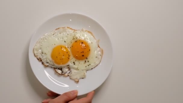 Two fried eggs on plate. Putting plate with fried eggs on wooden table. Close up white porcelain plate with fried egg. Traditional breakfast meal - Filmati, video