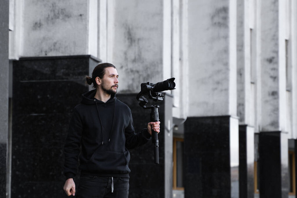 Young Professional videographer holding professional camera on 3-axis gimbal stabilizer. Pro equipment helps to make high quality video without shaking. Cameraman wearing black hoodie making a videos. - Zdjęcie, obraz