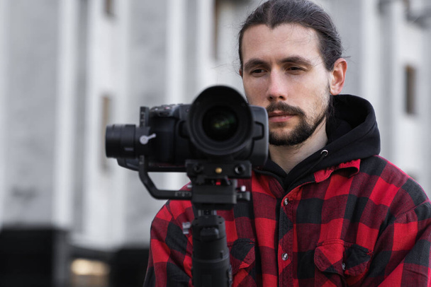Νέοι επαγγελματίες videographer κρατώντας επαγγελματική φωτογραφική μηχανή σε 3-άξονα gimbal σταθεροποιητή. Pro εξοπλισμός βοηθά να κάνει υψηλής ποιότητας βίντεο χωρίς ανακίνηση. Καμεραμάν φορώντας κόκκινο πουκάμισο κάνοντας βίντεο. - Φωτογραφία, εικόνα