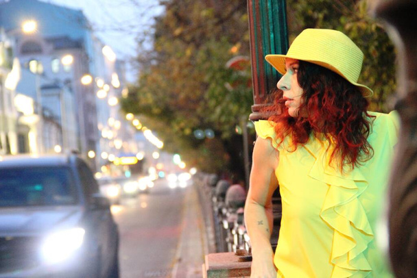 brunette lady stands on the road. A woman in a yellow hat and clothes against the background of the evening lights of the big city, passing car in the distance - Photo, Image