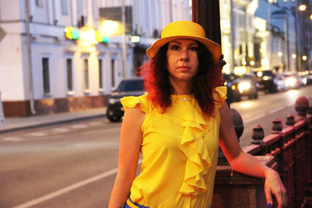 woman in a yellow hat and clothes against the background of the evening lights of the big city, passing car in the distance. brunette lady stands on the road. - Photo, Image