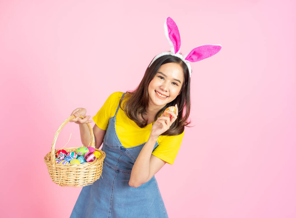 Half length studio shot of asian beauty happy young woman wearing bunny ears and holding colorful Easter egg in wood basket with lovely smile and colorful decor costume isolated on pastel backgrounds. - Photo, image