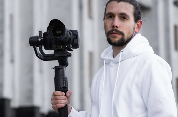 Fiatal profi videós professzionális kamerát tart 3 tengelyes gimbal stabilizátoron. Pro berendezések segít, hogy kiváló minőségű videót rázás nélkül. Az operatőr fehér kapucnis pulóvert visel, videókat készít. - Fotó, kép