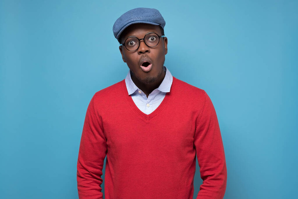 Beau noir afro-américain en pull rouge et chapeau bleu à l'air surpris. Studio tourné sur mur bleu
 - Photo, image