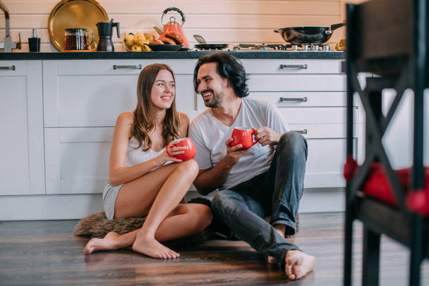 Young loving couple in the morning in the kitchen at home. A man and a woman are sitting on the floor laughing and having fun having breakfast in a beautiful stylish kitchen. - Fotó, kép
