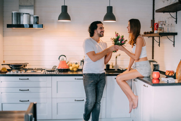 Kansainvälinen naistenpäivä. Nuori pari aamulla keittiössä kotona. Mies antaa naiselle kimpun tulppaaneja 8. maaliskuuta kauniissa tyylikkäässä keittiössä.. - Valokuva, kuva