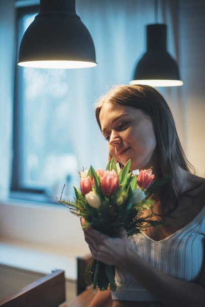 Portrait of a young woman with a bouquet of flowers. A beautiful girl sits on a table under the evening light of lamps with tulips in her hands. Sensual portrait in subdued light - Foto, Bild