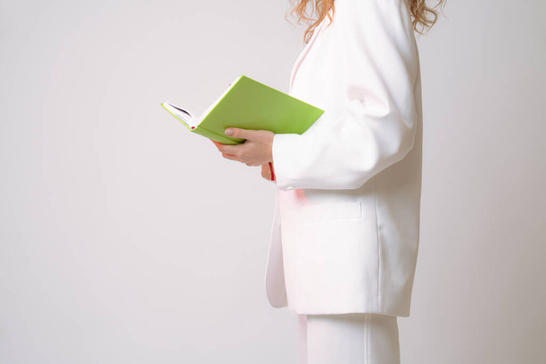 Boční pohled fotografie ženy v bílém obleku se zeleným zápisníkem v rukou, izolované přes šedé pozadí - Fotografie, Obrázek