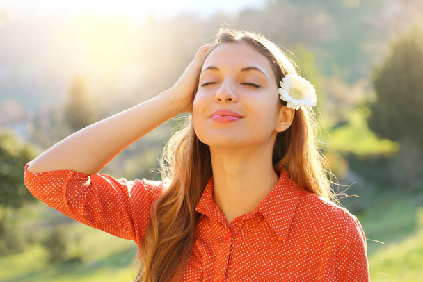 Spring girl. Close up portrait of beautiful relaxed young woman with white flower on ear breathing fresh air with closed eyes outdoor in spring time. - Photo, Image