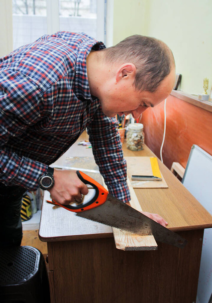 Ο ξυλουργός δουλεύει την τέχνη του σε ένα σκονισμένο εργαστήριο. Κοντινό πλάνο μιας νεαρής γυναίκας που χρησιμοποιεί πριόνι χειρός για να κόψει ξύλα σε ένα ξυλουργείο. - Φωτογραφία, εικόνα