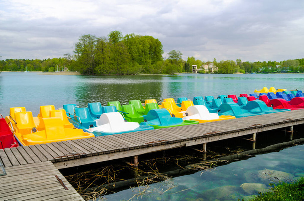 A wooden pier near lakeshore with the row of colored plastic catamarans with lake water and forest on the background. - Photo, Image