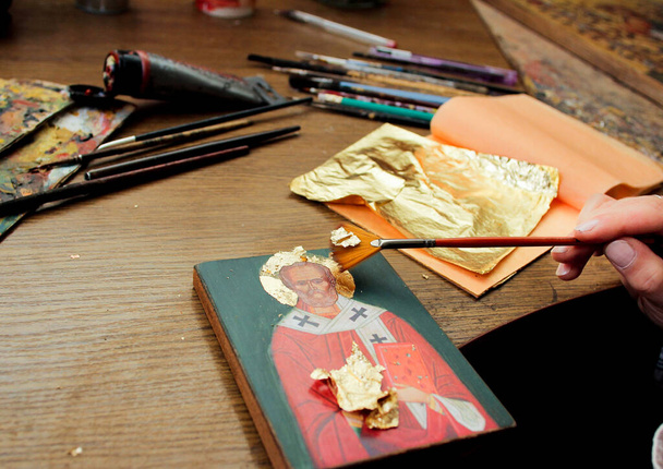 Χέρι με πινέλο από φύλλο χρυσού. Καλλιτέχνης εφαρμόζει φύλλο χρυσού στην εικόνα. Αναπαραγωγή της εικόνας του Αγίου Νικολάου. Η διαδικασία του έργου του καλλιτέχνη. Βούρτσες και μπογιές στο φόντο. - Φωτογραφία, εικόνα