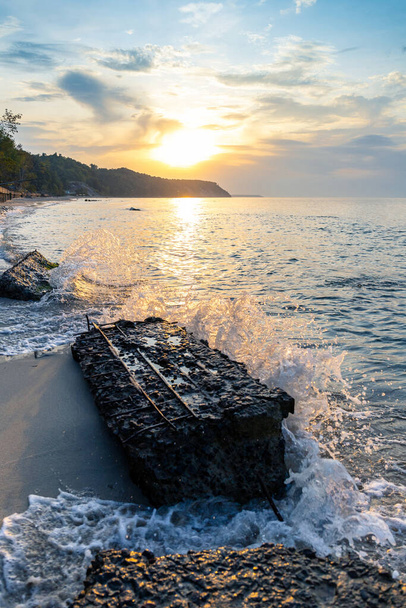 живописное побережье с лесистой скалой на заднем плане, низкое солнце во время заката и большие камни на переднем плане
 - Фото, изображение