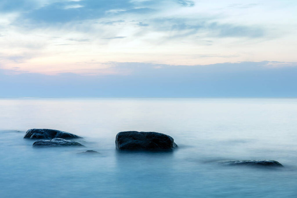 青い夕暮れに大きな石が水から突き出ている長い露出滑らかな海の表面 - 写真・画像