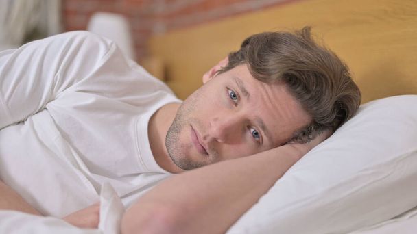 Portrét unaveného mladého muže při pohledu na kameru z postele - Fotografie, Obrázek