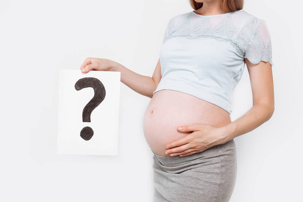 Kép egy terhes nőről, aki egy kérdőjellel ellátott papírt tart a terhes has közelében, fehér alapon - Fotó, kép