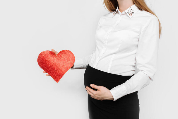 Image of pregnant business lady hugging her belly, holding red heart near her belly, on white background - Foto, Bild