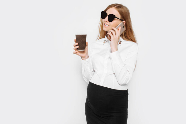 Egy fiatal, szemüveges terhes üzletasszony képe, egy lány, aki telefonon beszél és kávét iszik egy csészében, egy kis szünet a munkahelyén, fehér háttérrel. - Fotó, kép
