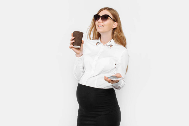 Image of a young pregnant businesswoman wearing glasses, a girl holding a smartphone and drinking coffee in a Cup, a break at work, on a white background - Photo, image