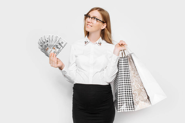 Obrázek mladé těhotné podnikatelky v brýlích pózování držení bankovek a tašky nakupování na bílém pozadí - Fotografie, Obrázek