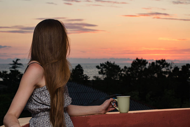 Elinde fincanla denize ve günbatımına bakan genç bir kadın. Yan görünüm. - Fotoğraf, Görsel