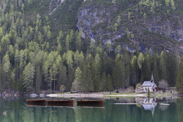 τοπίο του Lago di Braies, εντυπωσιακή πράσινη λίμνη, που αντανακλά τα ψηλά χιονισμένα βουνά, που περιβάλλεται από πλούσια βλάστηση, μπορούμε επίσης να δούμε μια ξύλινη καμπίνα που είναι η προβλήτα και μερικά ξύλινα σκάφη - Φωτογραφία, εικόνα