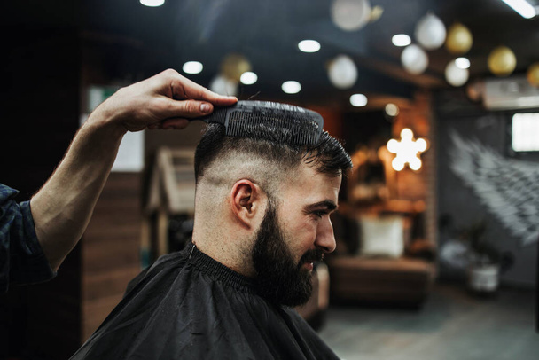 Good looking young adult man getting a hair and beard styling and dressing treatment by professional hairstylist. - Foto, Imagem