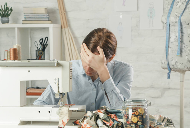 Sleepy, frustrated, exhausted seamstress with headache on her work desk. Small business or self-employed problems. Deadline or overwork concept, bored or getting tired at work in creative industry. - Photo, image