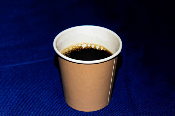 Egy közeli kép egy papírpohárról fekete kávéval az asztalon, amit a világoskék terítő borít. - Fotó, kép