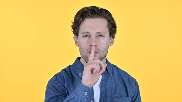 Fiatal férfi ujjal a szájon, csendes jel sárga háttér - Fotó, kép