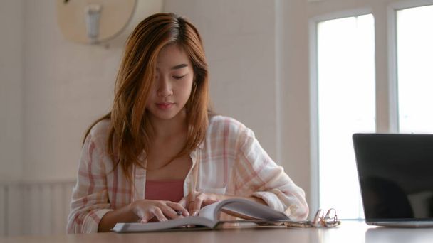 Nahaufnahme eines jungen College-Mädchens, das sich auf seine Aufgabe mit einem Buch auf dem Holztisch in der Bibliothek konzentriert - Foto, Bild