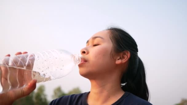 Азиатская молодая женщина пьет воду из пластиковых бутылок после пробежки в парке. Сбросить вес. Понятие здорового образа жизни
. - Кадры, видео