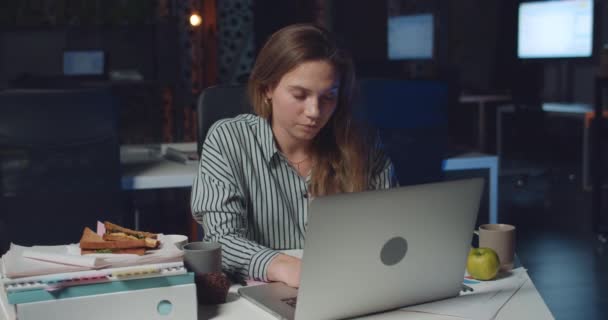 Front view of tired young woman typing on her laptop late at night. Stressed female employee using her personal computer in empty office . Concept of overwork and workaholism. - Filmati, video