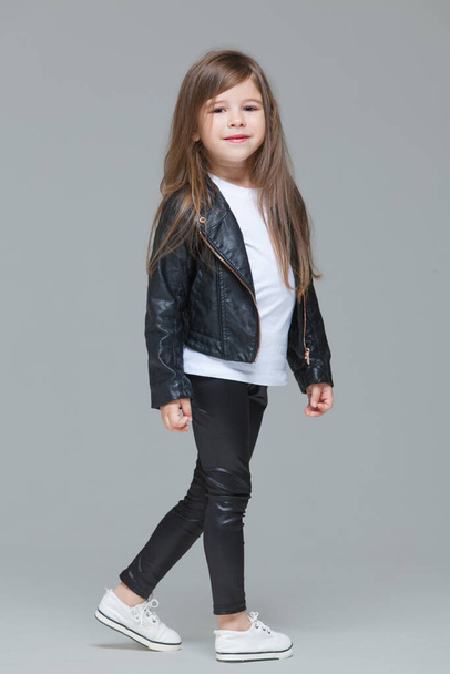 Baby girl with long hair in black leather jacket and leggings is standing in the studio on grey background - Photo, image