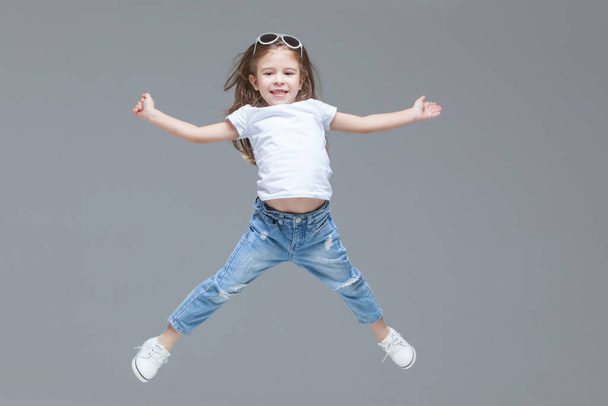 Kid girl preschooler in blue jeans, white t-shirt and sunglasses is jumping with raised hands isolated on grey background - Foto, Imagen