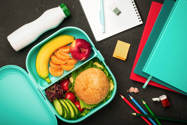 School lunch box with a hamburger and vegetables with fruits and school supplies on a dark background: the concept of a healthy and balanced diet. - Valokuva, kuva