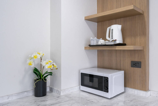Microwave cooking machine on white marble counter with electric kettle on wooden cabinet with yellow orchid flower in black ceramic vase interior kitchen decoration - Фото, зображення