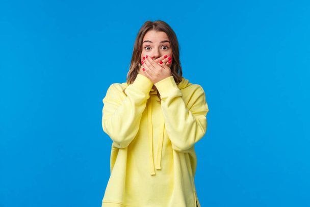 Shocked and speechless impressed blond woman close mouth with hands and stare astonished, hear shook rumour, gossiping make astounded face, standing blue background - 写真・画像