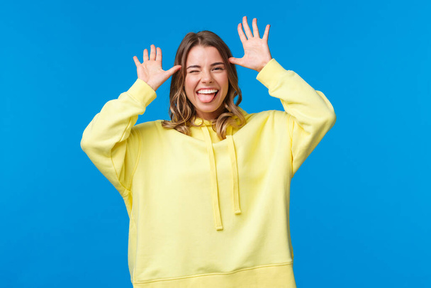 Portrait of alluring carefree caucasian blond girl having fun, showing catch me if you can, playful grimace with hands near head, stick tongue and smiling, standing blue background - Photo, Image