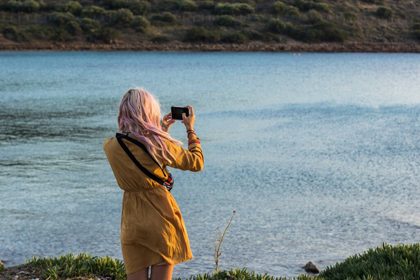 back to camera girl in yellow dress and small white travel bag take photo of tropic south nature landscape view in Mediterranean sea bey with boat and island in shot, evening time, soft colors - Photo, Image