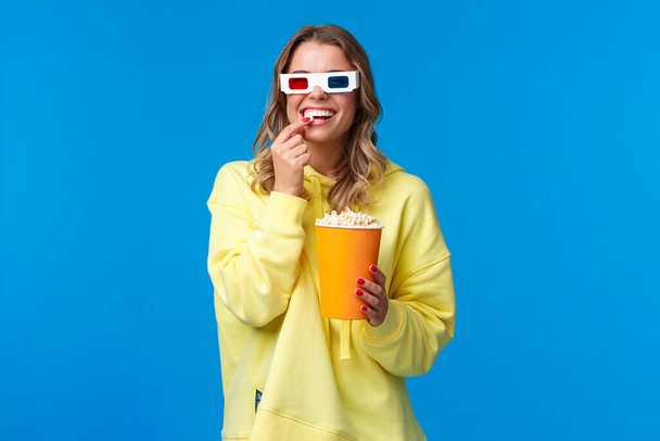 Leisure, fun and youth concept. Happy entertained smiling blond girl in 3d glasses, eating popcorn and laughing as watching funny comedy, go cinema watch movie with friends, blue backgorund - Zdjęcie, obraz
