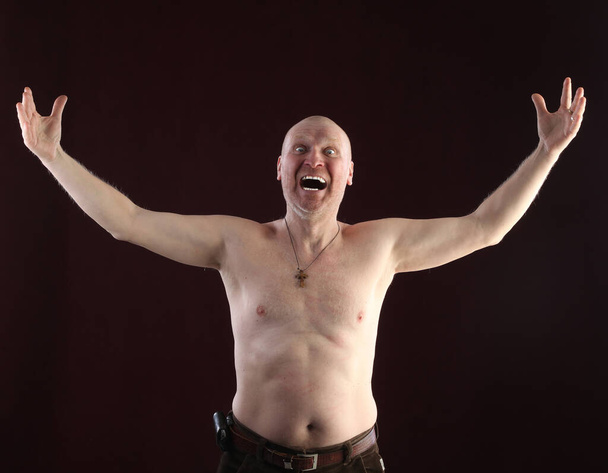 close-up portrait on a belt adult bald man with a naked torso and a cross on his chest on a dark background studio - Foto, Imagen