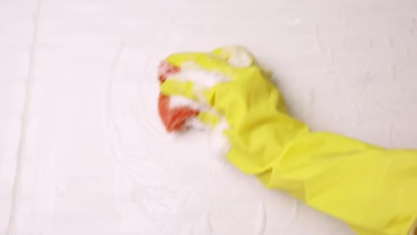 Een vrouwelijke hand in een gele latex handschoen sponst een tegel. Er ligt veel schuim in de gootsteen. Het concept van het schoonmaken van de badkamer, reinheid. Kopieerruimte. - Video