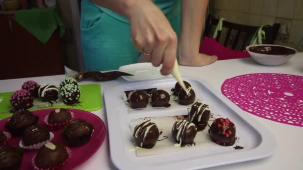 A woman decorates trickles of different chocolate balls from biscuit pulp in chocolate. Cooking Potato Cake. Next to the plates are cake blanks, liquid chocolate and colored sprinkles. - Séquence, vidéo