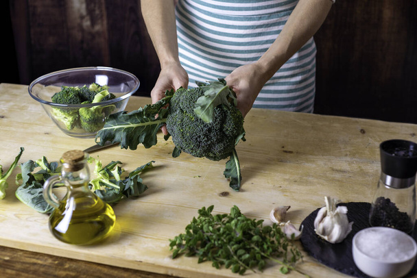 Moments of everyday life in Mediterranean cuisine: young female cook cleans broccoli on a wooden chopping board with extra virgin olive oil, garlic, pepper and knife on top - Photo, Image