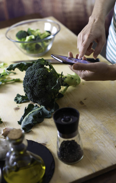 Moments of daily life in the Mediterranean cookery: young female cook consult her smartphone to find a recipe for broccoli on a wooden chopping board with extra virgin olive oil, pepper and a knife. - Photo, Image