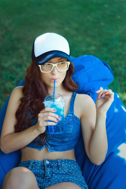 young woman of Caucasian appearance sitting on a puff on a lawn and having fun, makes emotions, she is in blue sportswear and a cap, summer vacation concept - Photo, Image