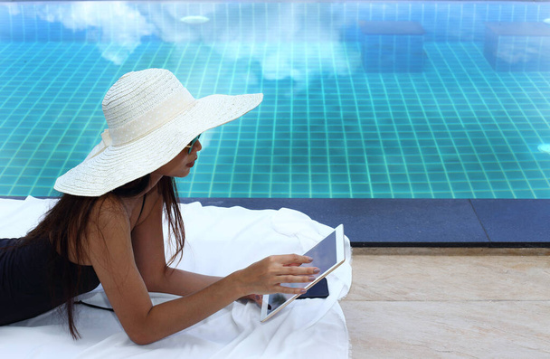 Along Side Pool lie with Mix Race Tanned Skin Woman in Big Hat black Swimsuit sunglasses tablet mobile phone iot take vacation holiday in Summer Island over blue Sky Cloud, copy space for text logo - Photo, Image