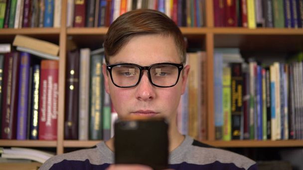 Um jovem está olhando para um smartphone em uma biblioteca. O homem com óculos olha cuidadosamente para o close-up do telefone. No fundo estão os livros sobre estantes. Biblioteca de livros
. - Foto, Imagem