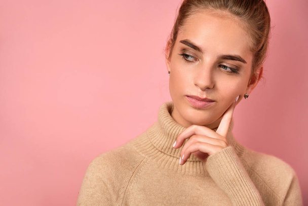 A girl with clean skin and a high bun stands on pink background and looks thoughtfully to the side, propping her cheek with finger - Foto, imagen